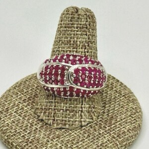 May include: A silver ring with a large oval-shaped setting filled with small red ruby stones. The ring is on a brown fabric ring display stand.