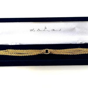 May include: Gold-toned bracelet with multiple strands, presented in a dark blue velvet box. The bracelet features an oval centerpiece and a clasp closure. The box is open, revealing the bracelet and the text "The Danbury Mint".