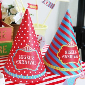 May include: Two party hats with a carnival theme. One is red with white polka dots, the other has red and blue stripes. Both hats feature a red label with the text "NIGEL'S CARNIVAL".