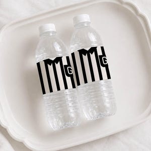 May include: Two clear plastic water bottles featuring custom labels with a black and white vertical striped pattern resembling a referee shirt, including a white collar graphic and a prominent letter G printed on the front of each label.