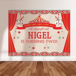 May include: Red and white circus themed birthday party backdrop with a red and white striped tent, a carousel horse, and the text "Come one and come all the magnificent Nigel is turning two!"