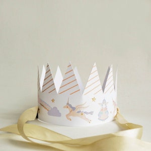 May include: A white paper crown with gold diagonal stripes and illustrations of unicorns, rainbows, and stars. The crown has a gold ribbon attached for securing. A whimsical party accessory.