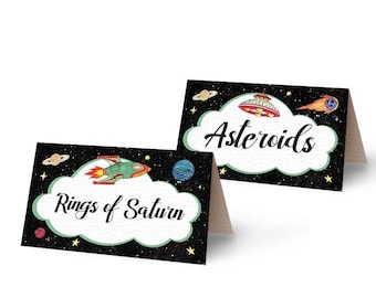 May include: Two black and white printable table cards with a space theme. The top card has a UFO and the text "Asteroids". The bottom card has a rocket ship and the text "Rings of Saturn".