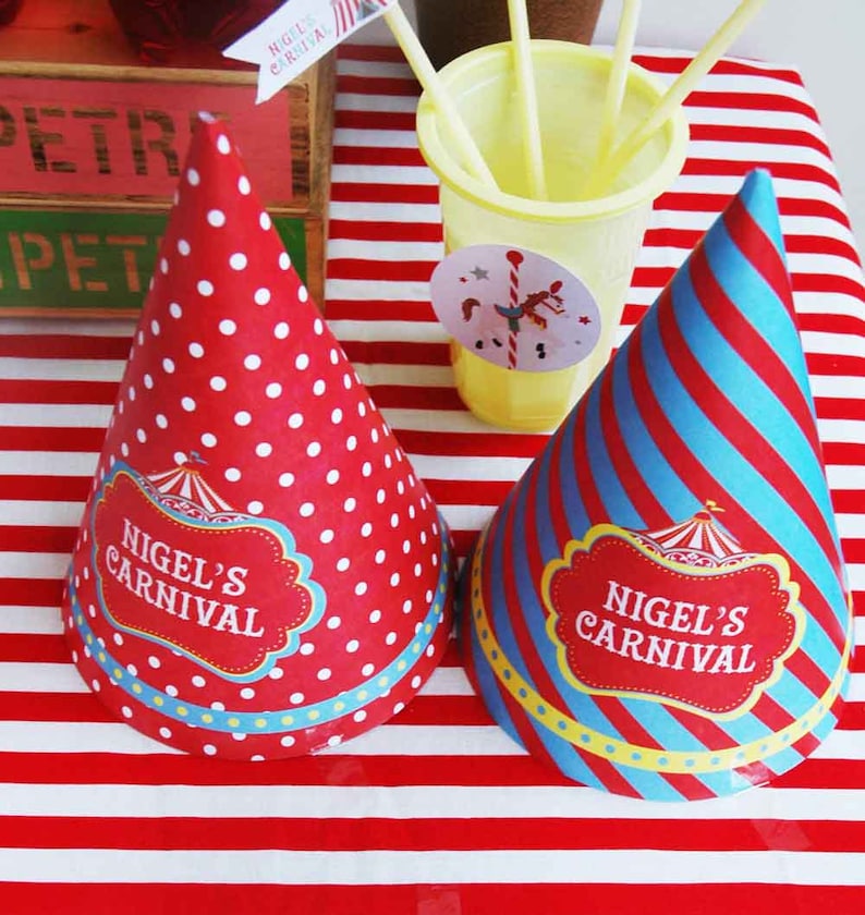 May include: Two party hats with "Nigel's Carnival" text. One hat is red with white polka dots, the other is blue and red striped. Both have a decorative label with a circus tent design. A yellow cup with straws and a small flag are also visible.