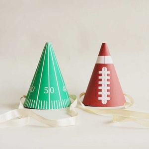 May include: Two party hats, one green with a football field design and the number 50, and one red with a football design. Both hats have white ribbon ties.