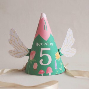 May include: A green and pink party hat with white wings and the text "Becca is 5" printed on the front.