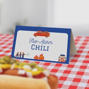May include: A blue and white table card with a red fire truck and firefighters. The text on the card reads "Five-Alarm Chili".