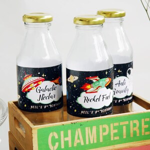 Puede incluir: Tres botellas de vidrio con etiquetas con un tema espacial. Las etiquetas tienen ilustraciones de un cohete, un OVNI y un astronauta. Las etiquetas también incluyen el texto "Galactic Nectar", "Rocket Fuel" y "Anti Gravity".