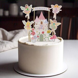 May include: A white frosted cake decorated with a fairy-themed cake topper. The topper features a castle, fairies, flowers, and the name "Rebecca" on a banner. The number "2" is also displayed.