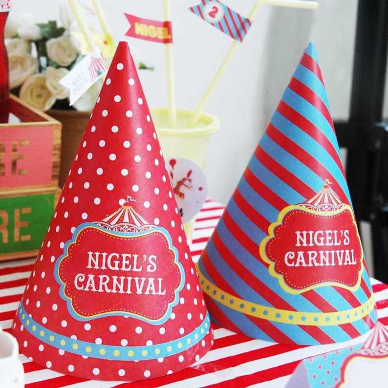 May include: Two party hats with a carnival theme. One is red with white polka dots, the other has red and blue stripes. Both hats feature a red label with the text "NIGEL'S CARNIVAL".