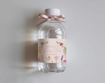 Peut inclure: Une bouteille d'eau en plastique transparente avec un ruban rose et une étiquette personnalisée qui dit "Becca's Fairy First Birthday". L'étiquette présente un design fantaisiste avec des fées, des fleurs et un fond rose clair.