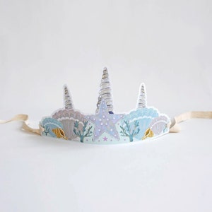 May include: A paper crown with a blue, white, and gold seashell design. The crown features a large, white starfish with pink and blue accents. The crown is tied with a white ribbon.