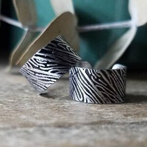 May include: Two silver rings with a textured, zebra-like pattern. The rings are wide bands, and the design is a contrast of black and silver. The rings are displayed on a wooden surface, with a blurred background of green and white.