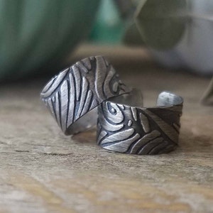 May include: Two silver ear cuffs with a textured, abstract design. The cuffs are made of metal and have a brushed finish.