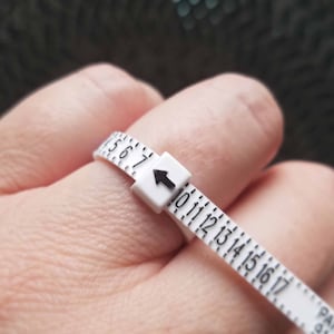 Peut inclure: Un mesureur de bagues blanc avec des chiffres noirs et un indicateur de fl&egrave;che est enroul&eacute; autour d'un doigt. Le mesureur mesure les tailles de bagues de 5 &agrave; 17. L'arri&egrave;re-plan est flou.