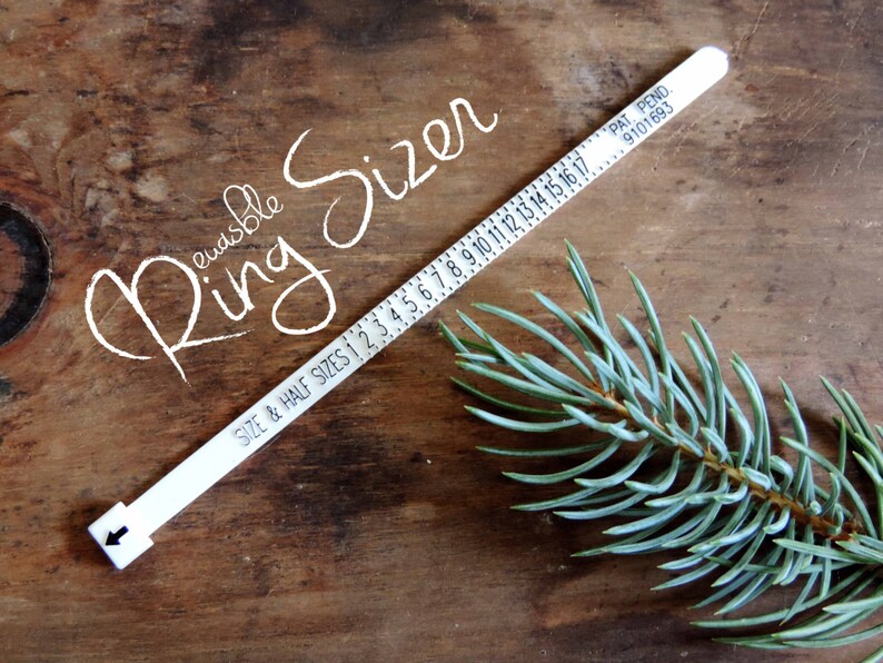 Peut inclure: Un baguier r&eacute;utilisable blanc avec des marquages noirs et le texte "SIZE & HALF SIZES". Le baguier est pos&eacute; sur une surface en bois avec une branche d'aiguilles de pin bleu-vert. Le texte "Reusable Ring Sizer" est &eacute;crit en cursive.