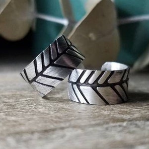 May include: Two silver ear cuffs with a geometric leaf pattern. The cuffs are made of metal and have a brushed finish.