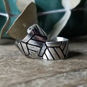 May include: Two silver rings with a geometric design. The rings have a textured surface and a wide band. The design is made up of a series of angled lines that create a pattern. The rings are on a wooden surface.