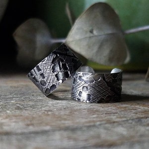 May include: Two silver rings with a textured floral design. The rings are on a wooden surface.