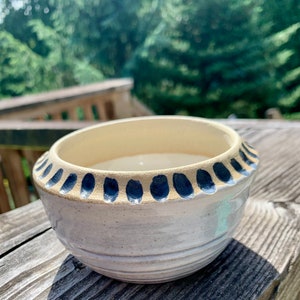 May include: A ceramic bowl with a cream-coloured interior and a white exterior featuring horizontal ridges. The rim is decorated with a repeating pattern of dark blue, oval-shaped indentations that create a textured border around the top edge.