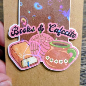 May include: A sticker featuring the words "Books & Cafecito" with illustrations of a book, a concha pastry, and a coffee mug. The sticker has a brown background with holographic accents and is held by a hand.