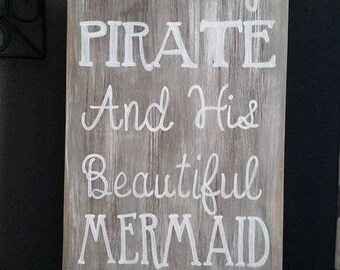 May include: A rustic wooden sign with white painted text that reads "A Salty Pirate And His Beautiful Mermaid Bathe Here".