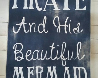 May include: A rectangular wooden sign with a navy blue background and white lettering. The text reads "A Salty Pirate And His Beautiful Mermaid Bathe Here" in a whimsical font, perfect for a nautical-themed bathroom or beach house.