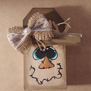 May include: A wooden scarecrow decoration with a painted face, blue eyes, and an orange nose. A burlap bow with white lace and a pearl accent adorns the top. The scarecrow has a wooden hat and a painted smile.