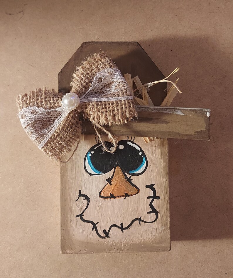 May include: A wooden scarecrow decoration with a painted face, blue eyes, and an orange nose. A burlap bow with white lace and a pearl accent adorns the top. The scarecrow has a wooden hat and a painted smile.