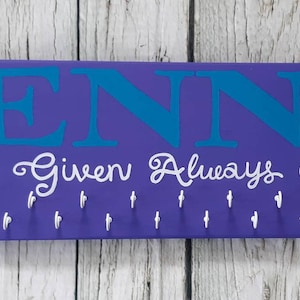 May include: Purple wooden medal display with the name "JENNA" in teal blue lettering and the phrase "Never Given Always Earned" in white cursive lettering. The display has 20 hooks for hanging medals.