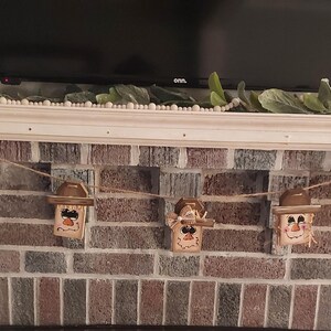 May include: A garland of wooden scarecrow decorations, each with a painted face, straw hair, and a brown hat. The scarecrows are strung on twine and displayed against a brick fireplace, ideal for autumn decoration.