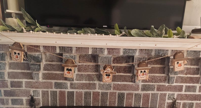 May include: A garland of wooden scarecrow decorations, each with a painted face, straw hair, and a brown hat. The scarecrows are strung on twine and displayed against a brick fireplace, ideal for autumn decoration.