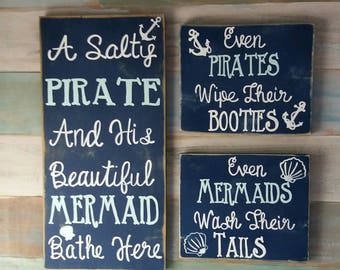 May include: Three wooden signs with nautical themes. The largest sign is blue with white text that reads "A Salty Pirate And His Beautiful Mermaid Bathe Here". The smaller signs are also blue with white text and nautical symbols. One sign reads "Even Pirates Wipe Their Booties" and the other reads "Even Mermaids Wash Their Tails".