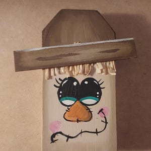 May include: Hand-painted wooden wall decoration with a cartoon face. It has large black eyes, an orange beak, and pink cheeks. The design includes a brown hat and straw-like fringe, creating a rustic, folk art aesthetic.
