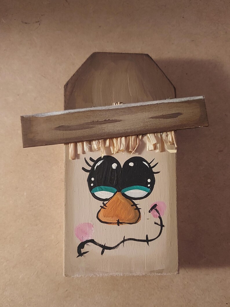 May include: Hand-painted wooden wall decoration with a cartoon face. It has large black eyes, an orange beak, and pink cheeks. The design includes a brown hat and straw-like fringe, creating a rustic, folk art aesthetic.