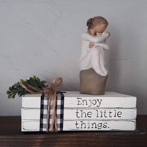 May include: A stack of three white books with black and white checkered ribbon. The books are stacked on a wooden surface. The top book has the text "Enjoy the little things." A white porcelain figurine of a woman holding a child is on top of the books.