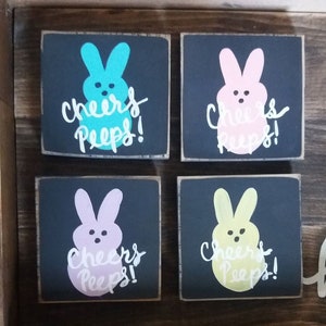 May include: Four wooden signs with black backgrounds and white lettering. Each sign features a different colored Peep, a marshmallow candy shaped like a chick, with the text "Cheers Peeps!"
