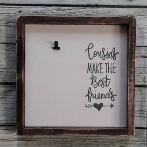 May include: A wooden framed sign with a white background and black text that reads "Cousins make the best friends" with a small black heart and arrows. A black clip is attached to the top of the sign.
