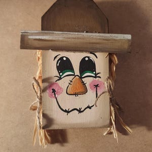 May include: A wooden scarecrow face with a painted smile, rosy cheeks, and an orange nose. The eyes are black with green irises. The scarecrow has a brown hat and straw hair tied with twine. A charming autumn decoration.