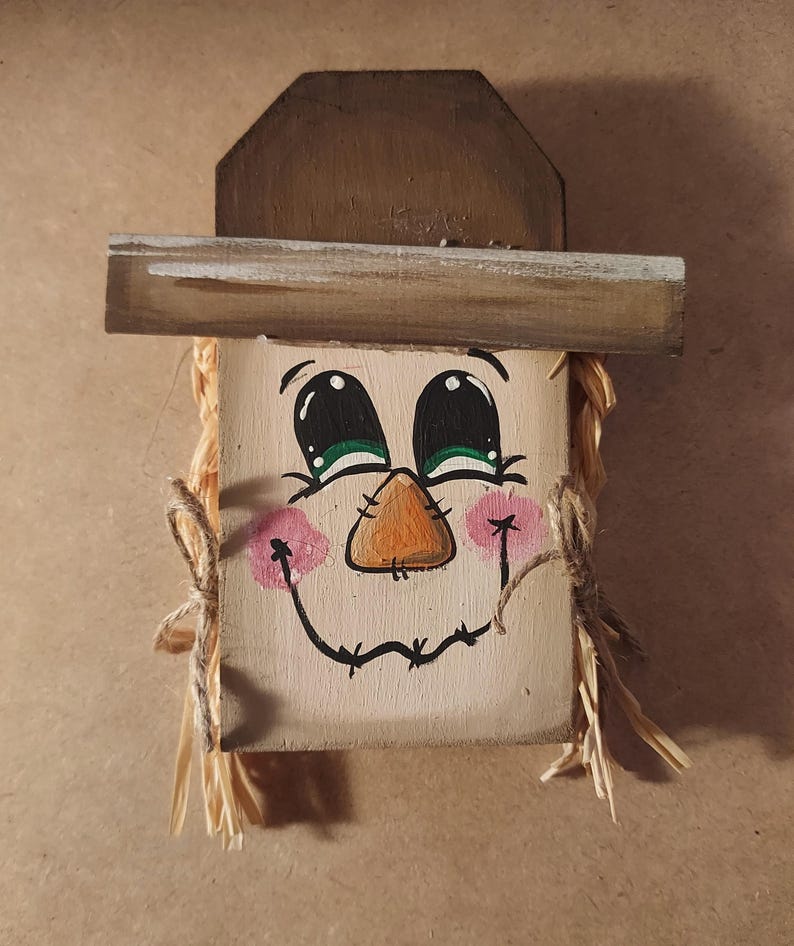 May include: A wooden scarecrow face with a painted smile, rosy cheeks, and an orange nose. The eyes are black with green irises. The scarecrow has a brown hat and straw hair tied with twine. A charming autumn decoration.