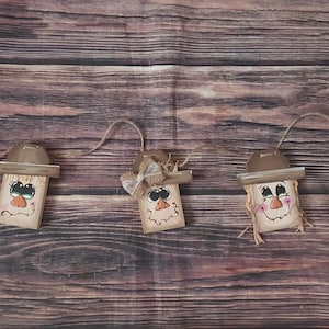 May include: A garland of five wooden scarecrow decorations. Each scarecrow features a painted face, a brown hat, and straw hair. The scarecrows are strung together with twine, ideal for autumn decoration.