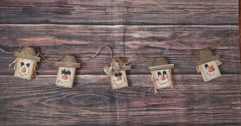 May include: A garland of five wooden scarecrow decorations. Each scarecrow features a painted face, a brown hat, and straw hair. The scarecrows are strung together with twine, ideal for autumn decoration.