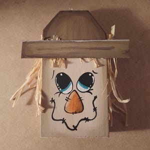 May include: A painted wooden scarecrow face with a brown hat and straw hair. The face features large blue eyes, an orange nose, and a stitched smile. Ideal for autumn decorations and seasonal displays.