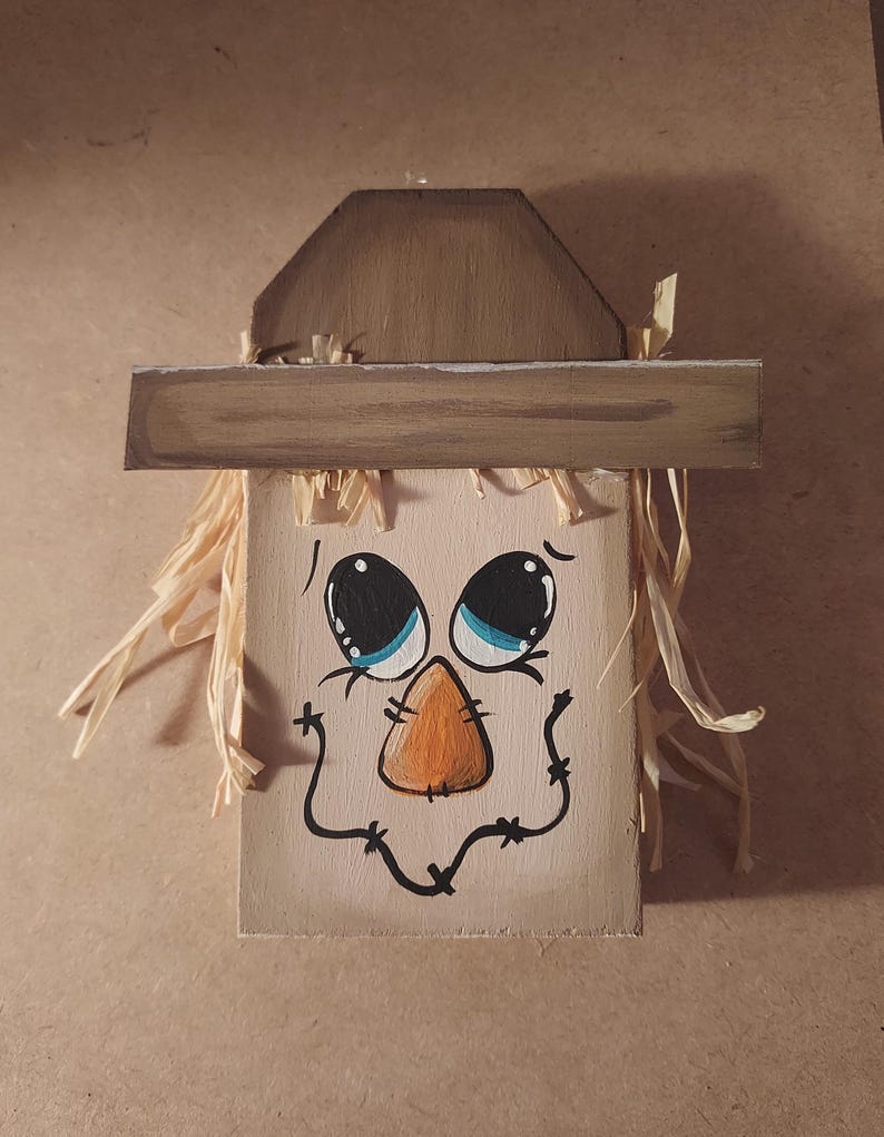 May include: A painted wooden scarecrow face with a brown hat and straw hair. The face features large blue eyes, an orange nose, and a stitched smile. Ideal for autumn decorations and seasonal displays.