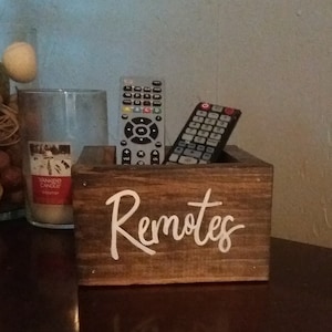 May include: A wooden box with the word "Remotes" written on the front in white paint. Two remote controls are inside the box.