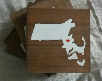 May include: Four wooden coasters with a white painted outline of the state of Massachusetts. The state of Massachusetts is painted white with a small red heart in the center of the state.