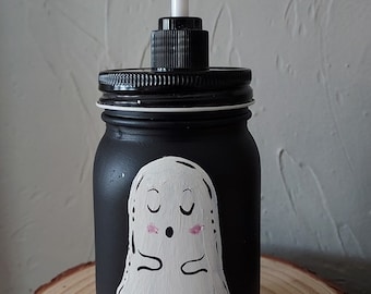 Peut inclure: Distributeur de savon en pot Mason noir avec une pompe noire. Le pot est peint avec un motif de fantôme blanc avec des joues roses. Le distributeur est posé sur une tranche de bois.
