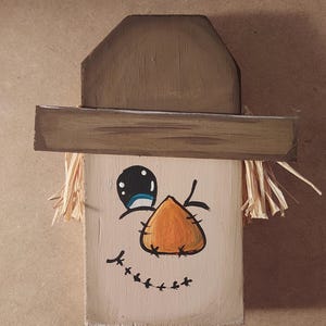 May include: A wooden scarecrow face with a painted orange nose, black eyes, and a stitched smile. The scarecrow has a brown hat and straw hair. The face is painted on a light beige wooden block, ideal for autumn decorations.