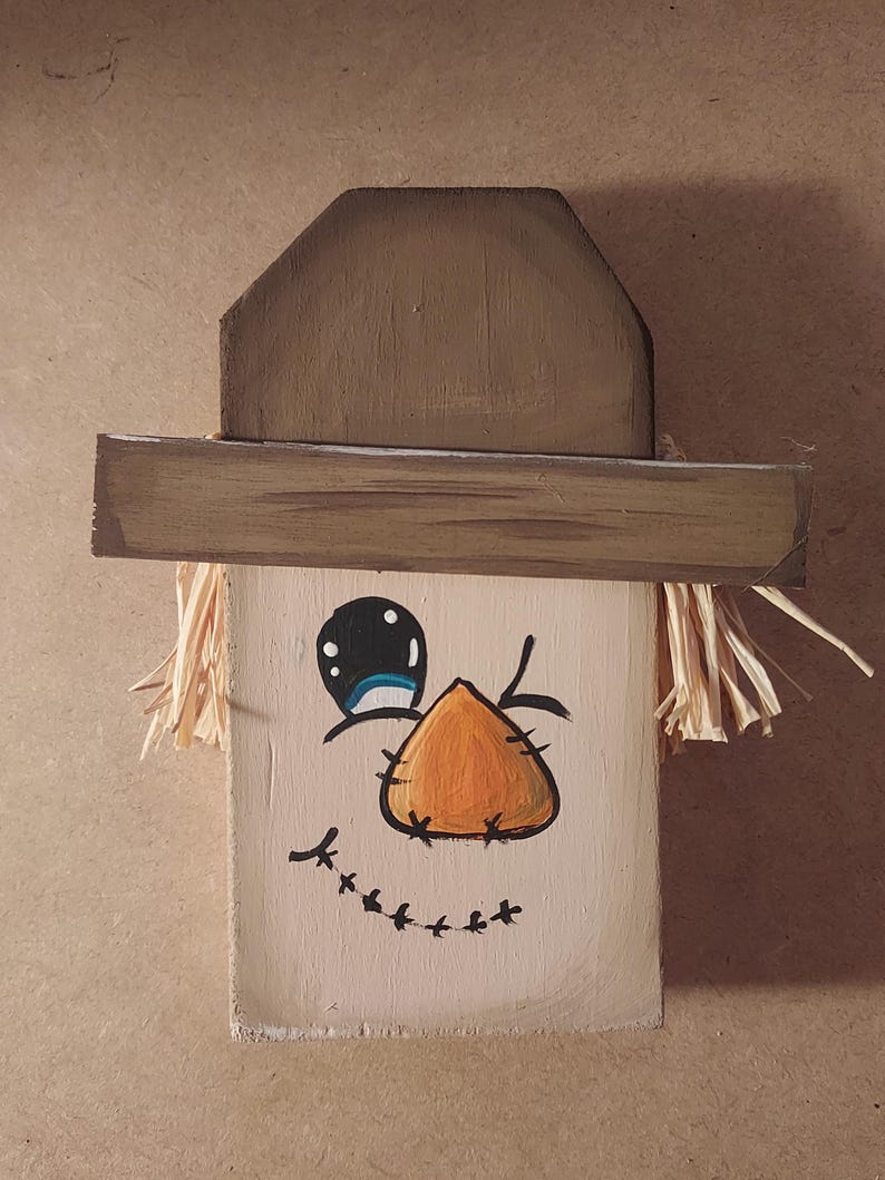 May include: A wooden scarecrow face with a painted orange nose, black eyes, and a stitched smile. The scarecrow has a brown hat and straw hair. The face is painted on a light beige wooden block, ideal for autumn decorations.