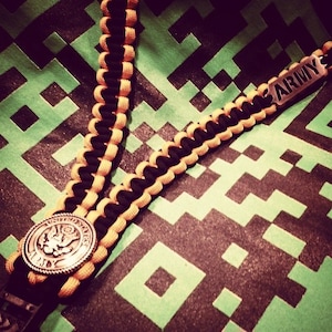 May include: A black and yellow paracord lanyard with a silver metal charm that says "ARMY". The lanyard is on a green and black patterned background.